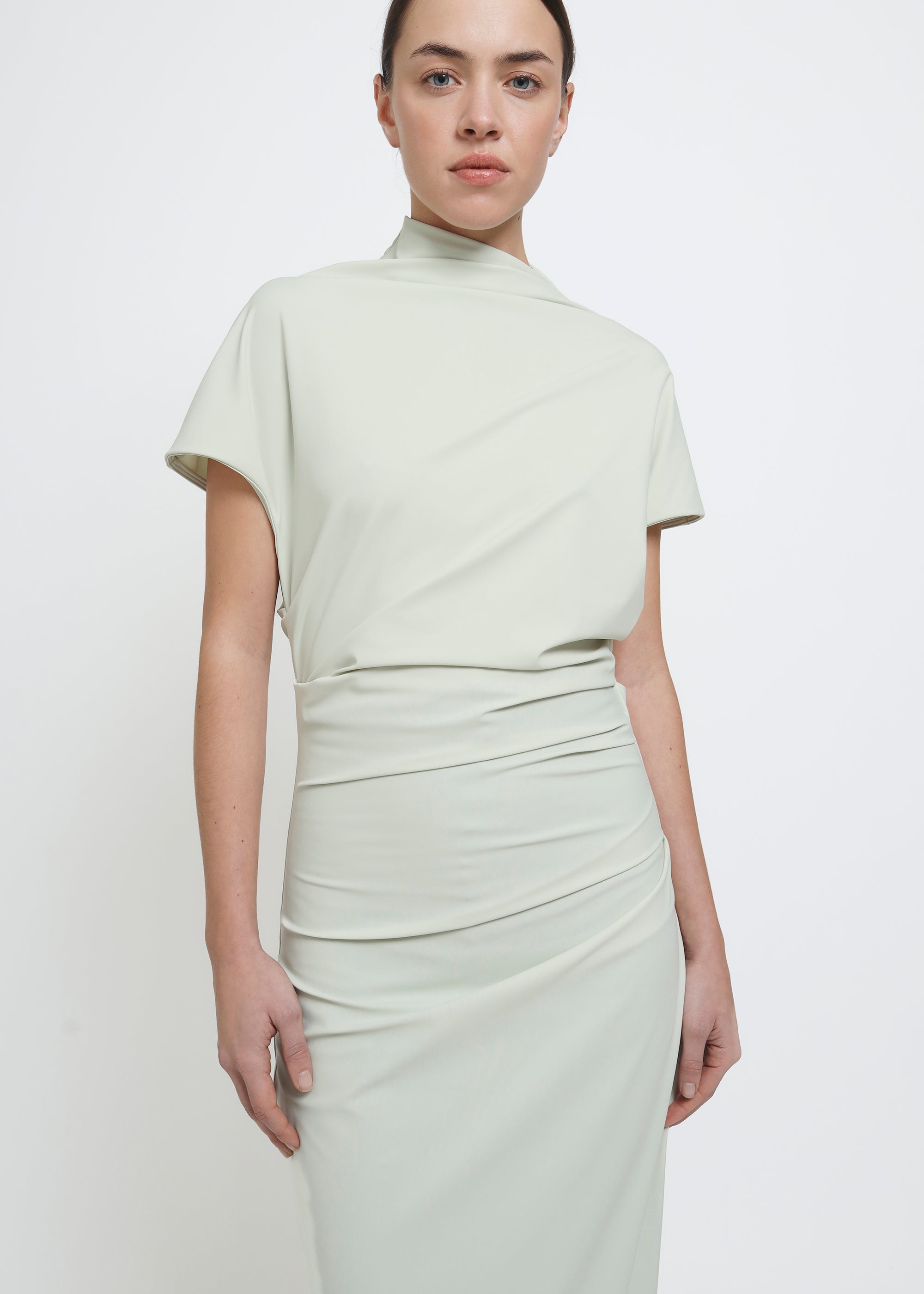 A model wearing a light sage green mock-neck short-sleeve dress with gathered draping details at the waist and hips.