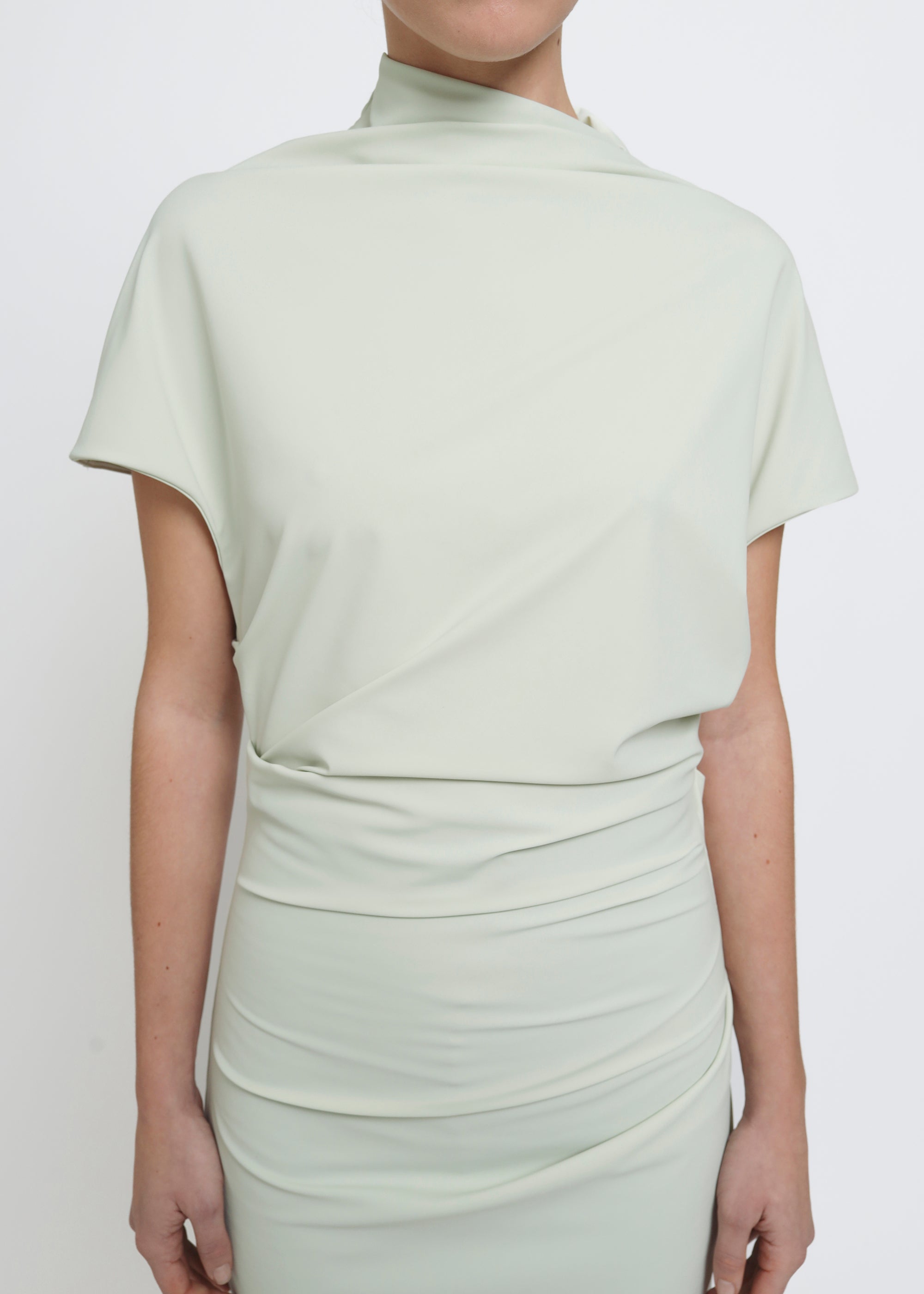 A model wearing a light sage green mock-neck short-sleeve dress with gathered draping details at the waist and hips.