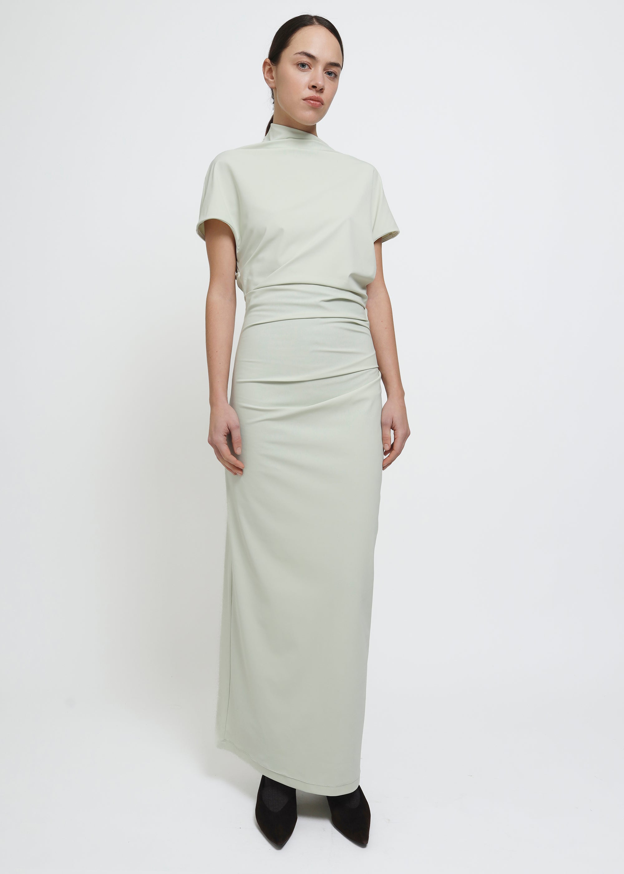 A model wearing a light sage green mock-neck short-sleeve dress with gathered draping details at the waist and hips.