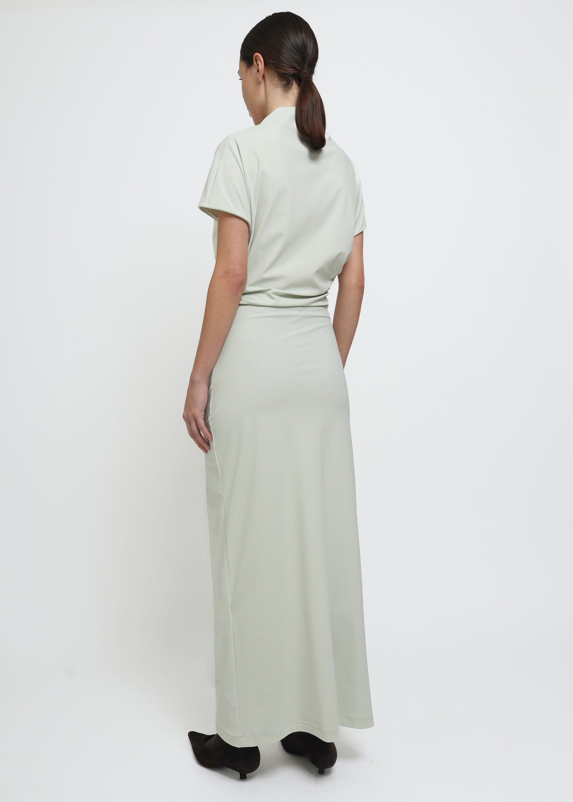 A model wearing a light sage green mock-neck short-sleeve dress with gathered draping details at the waist and hips.